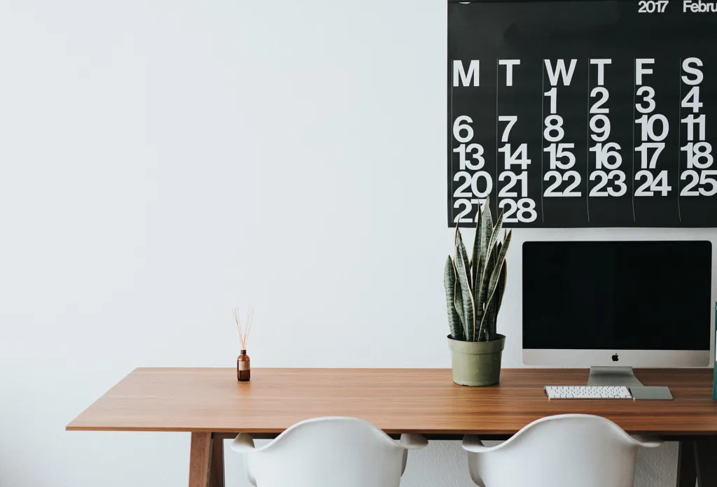 Clean, minimal wooden desk with a computer, plant and white chairs in a bright room