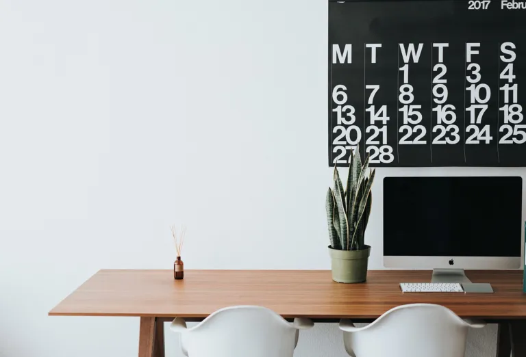 Clean, minimal wooden desk with a computer, plant and white chairs in a bright room