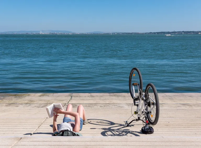 Person relaxing and reading a book on a waterfront pier beside a bicycle on a sunny day