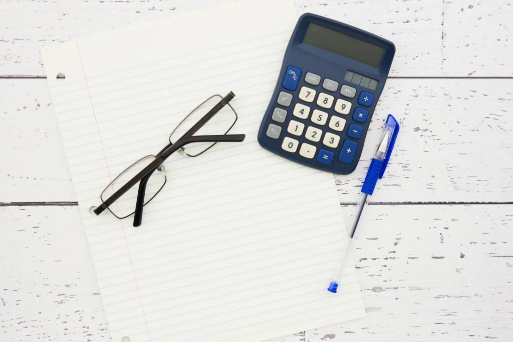 Calculator, lined paper, pen and glasses on a white wooden desk