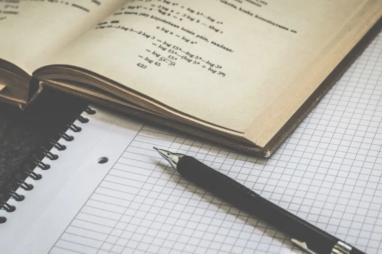 Open math textbook with equations beside a grid notebook and pen